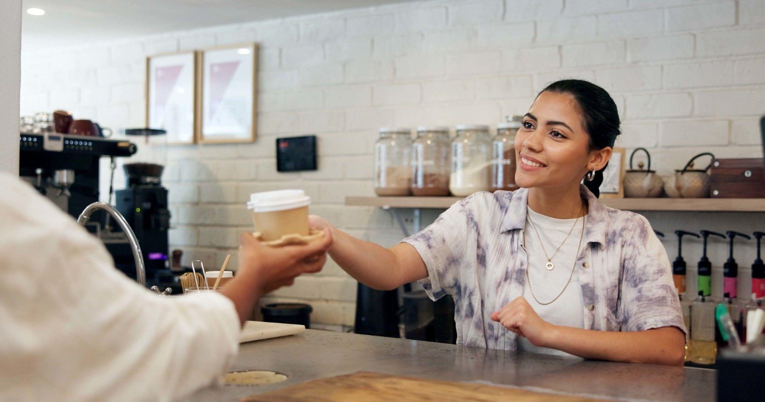 Smiling café employee handing a takeaway coffee to a customer across the counter